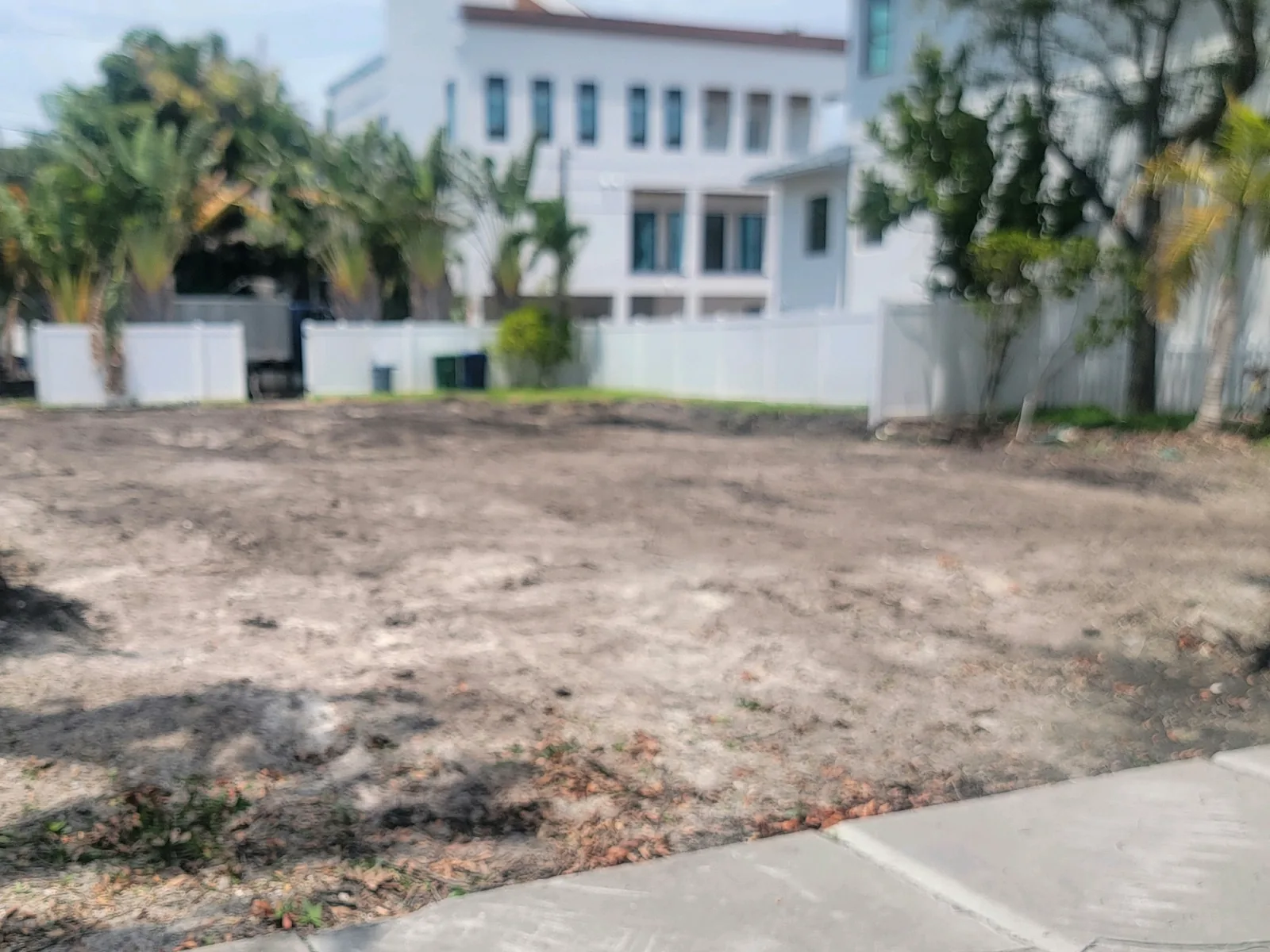 Cleared dirt lot after a house demolition, with a sidewalk in front and neighboring homes behind