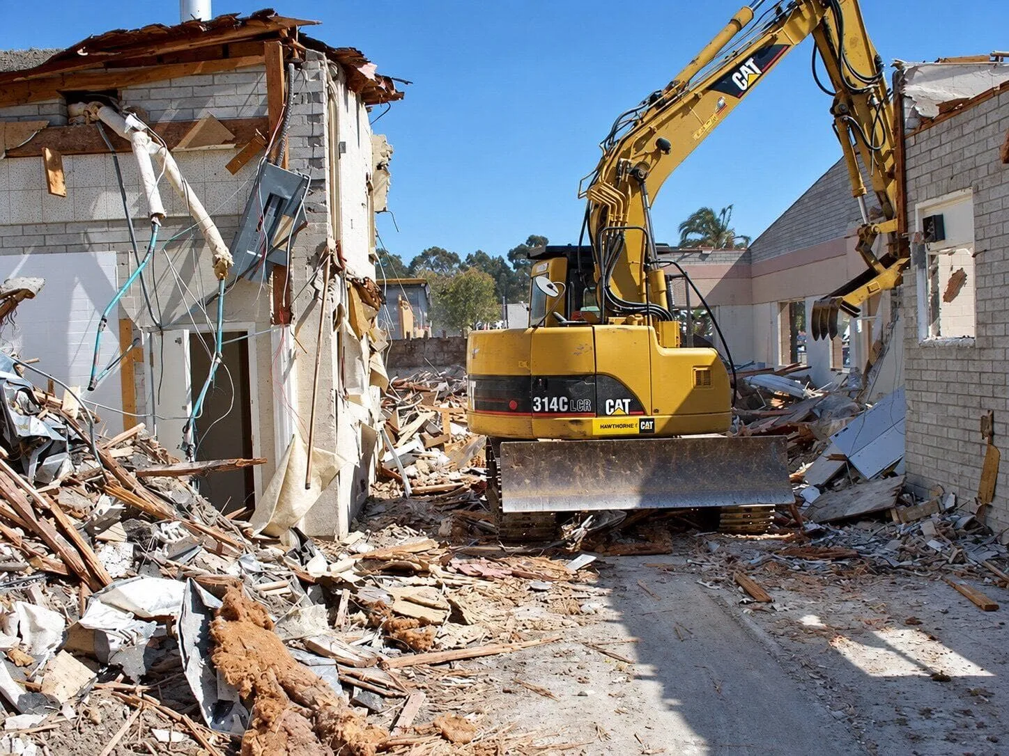 Excavator working through a gutted block commercial building, with roof framing, wiring and debris piled on both sides