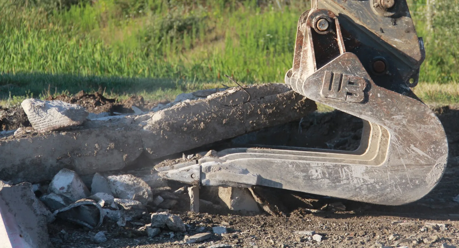 Excavator bucket and thumb lifting a broken section of concrete slab out of the ground during concrete removal