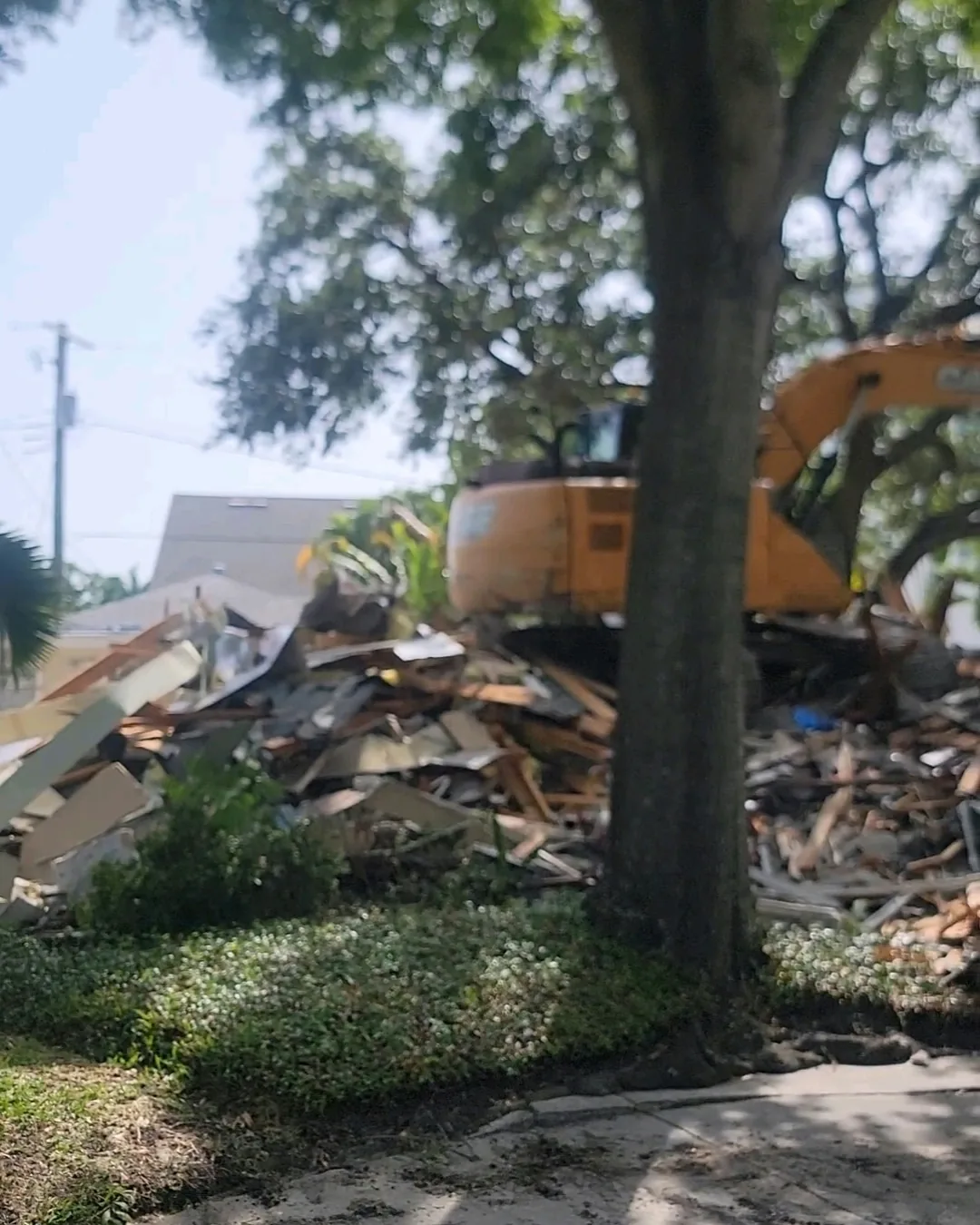Pile of demolition debris with an excavator parked behind a tree at the edge of the lot