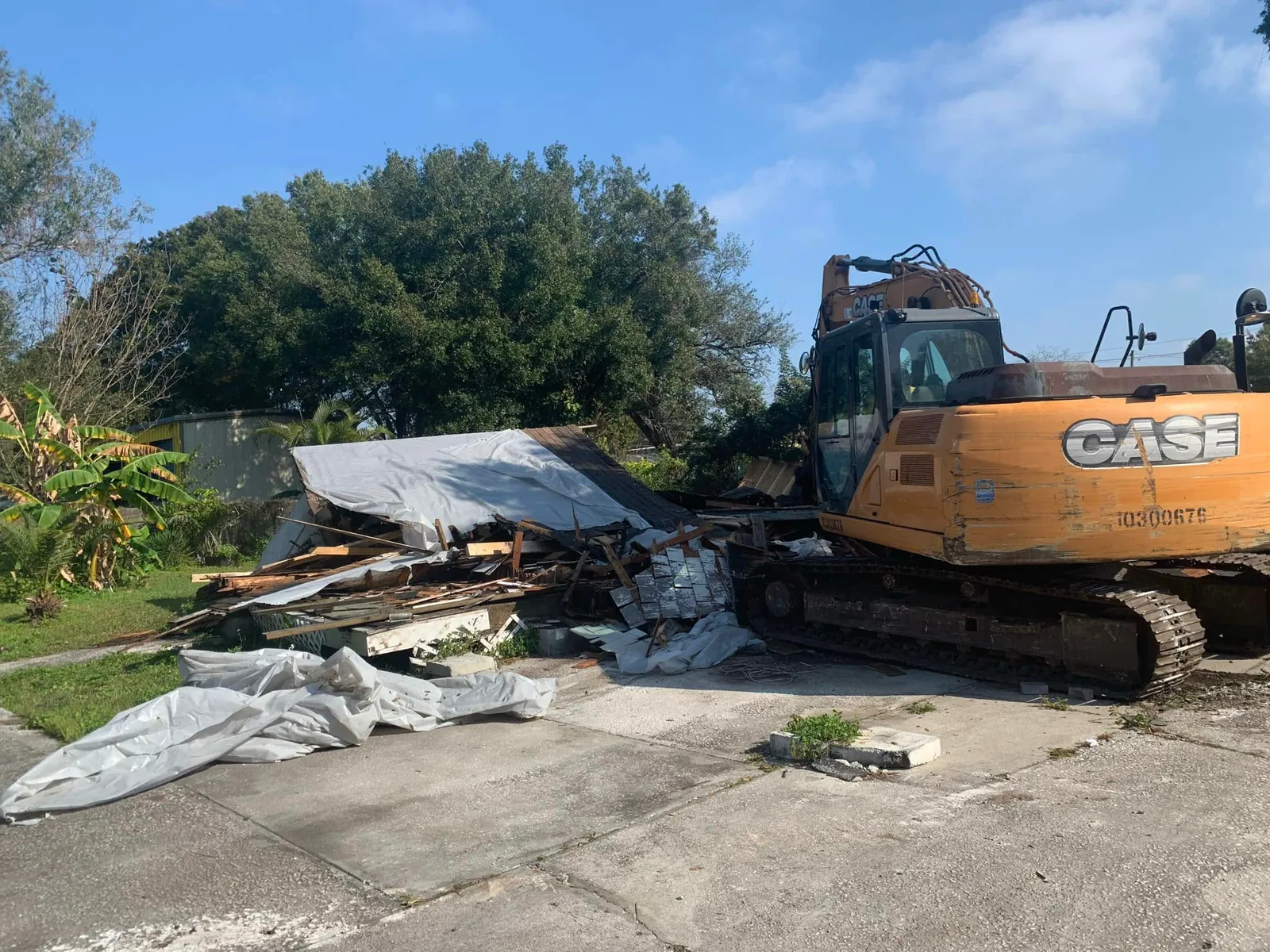 Excavator crushing a small backyard outbuilding, with tarps and loose lumber on the ground