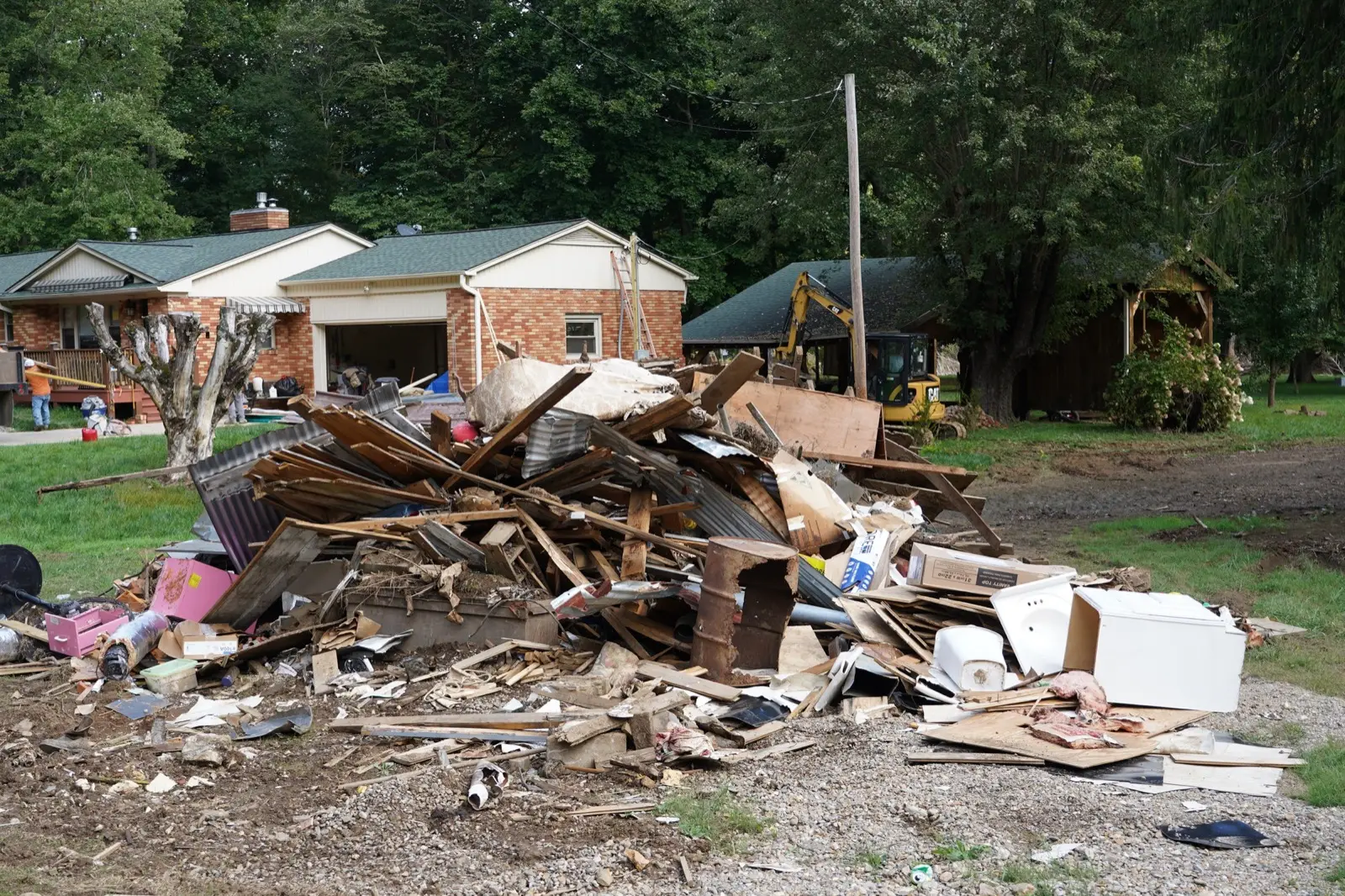 Pile of flood-soaked lumber, drywall and household contents in front of a brick ranch house, with an excavator working behind it