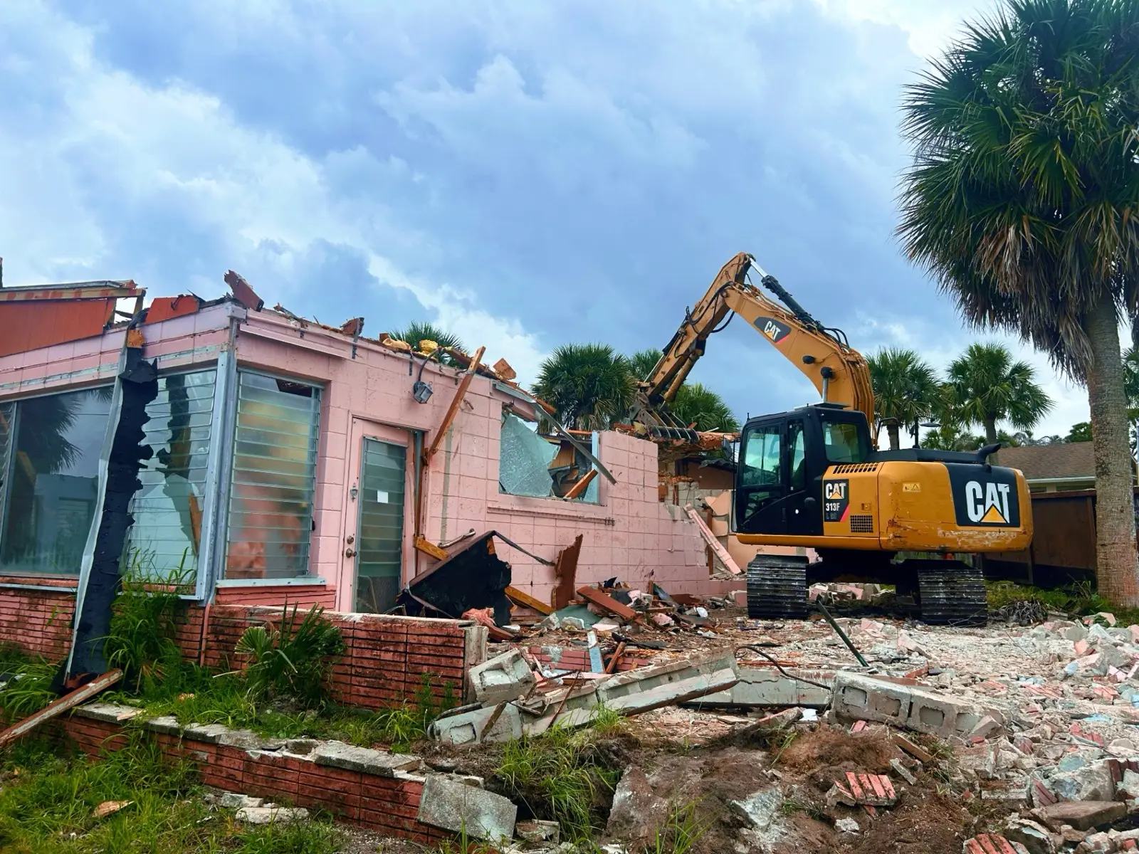 Excavator tearing down an older pink block house with jalousie windows, with palm trees behind it