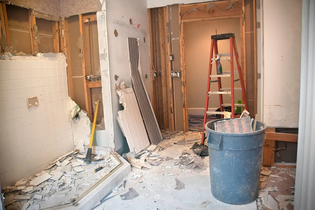 Bathroom gutted to the studs during interior demolition, with broken wall tile on the floor and a debris can beside a ladder