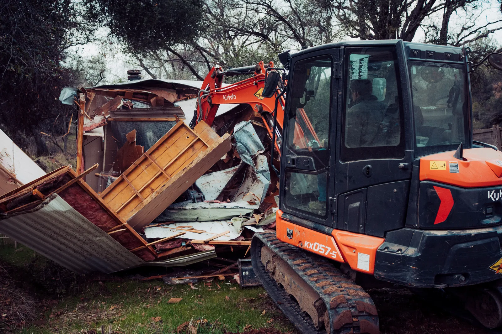 Excavator tearing apart a mobile home, pulling the roof and wall panels down into a debris pile
