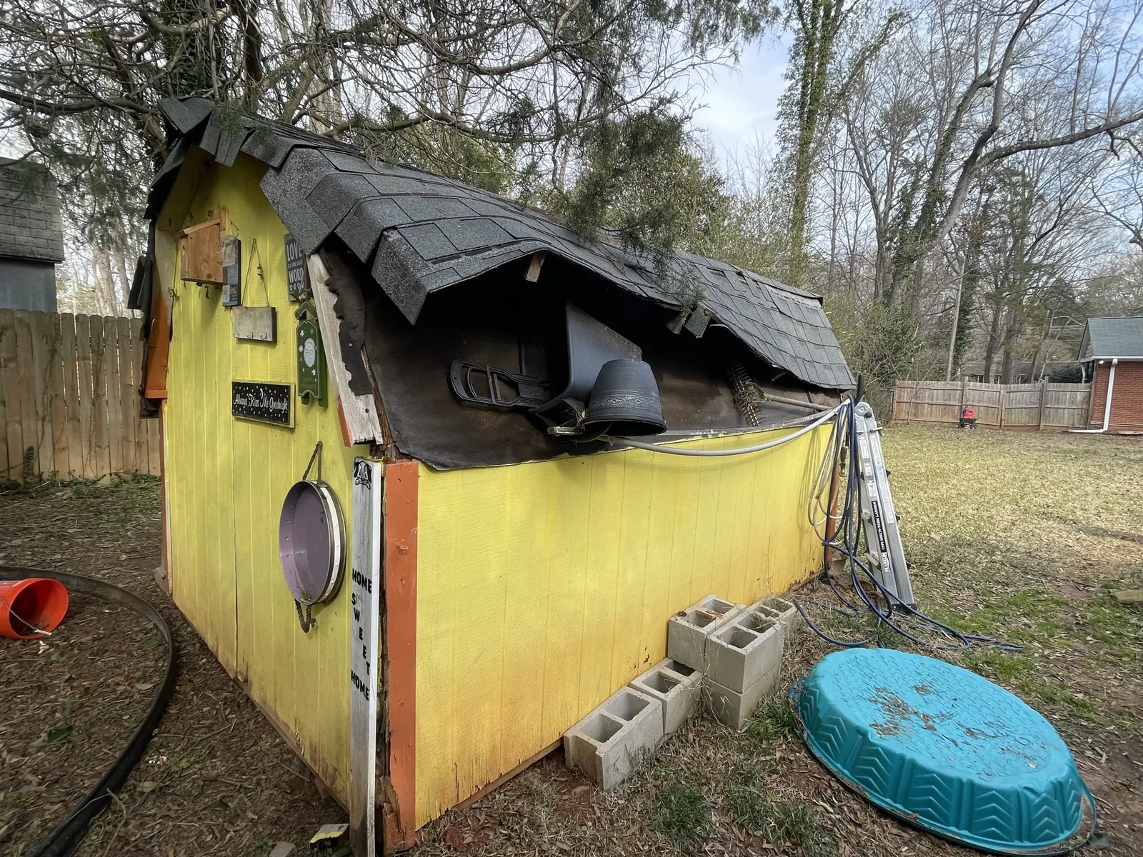 Old yellow backyard shed with a sagging, caved-in roof and concrete blocks stacked against its side, waiting for removal