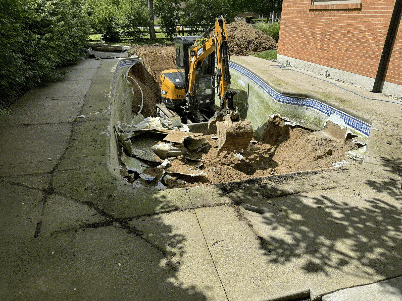 Compact excavator working inside a drained inground pool, breaking out the shell walls and liner during a pool removal