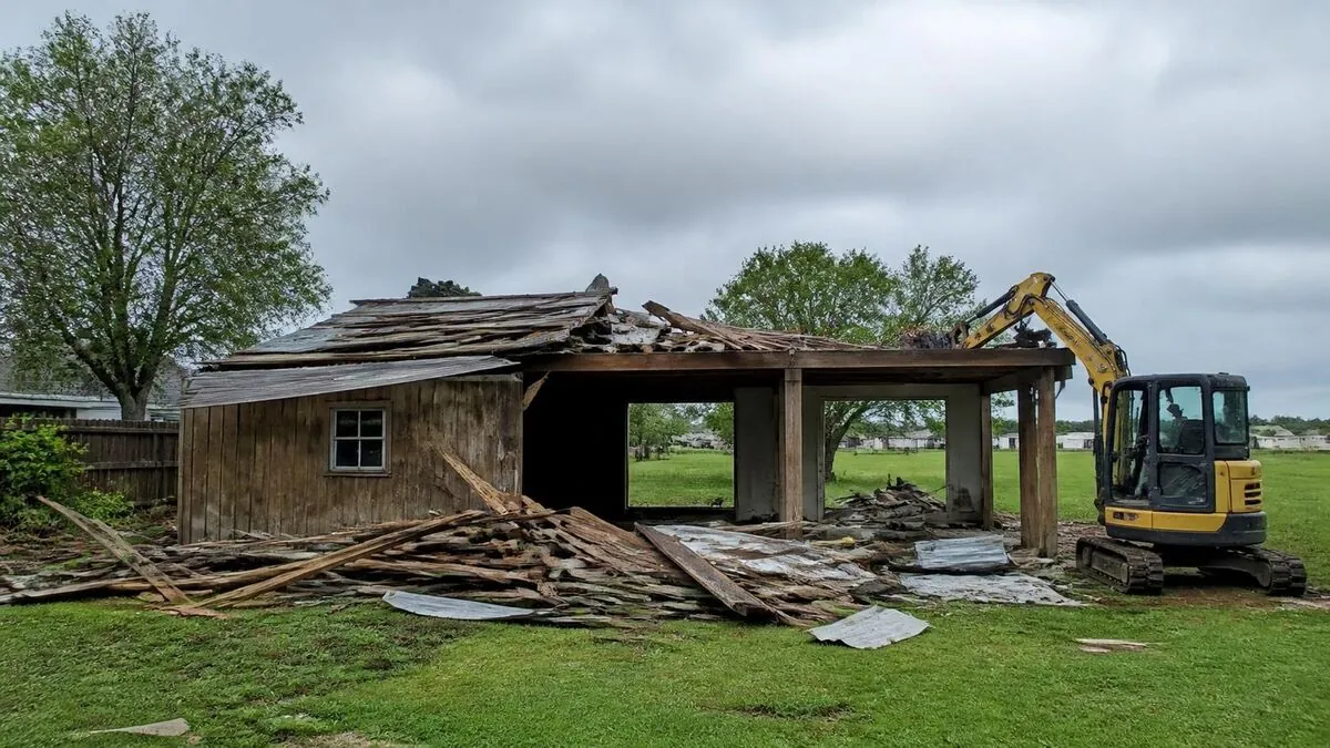 Shed demolition in progress: an excavator tearing down an old wood outbuilding, with broken boards and roofing tin piled on the grass